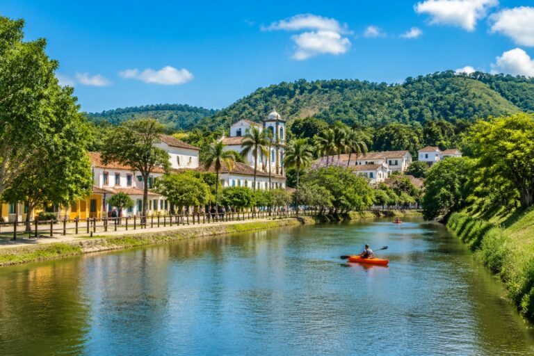 Vista de Guararema com casas coloridas, colinas verdes, rio tranquilo e pessoas aproveitando atividades ao ar livre.