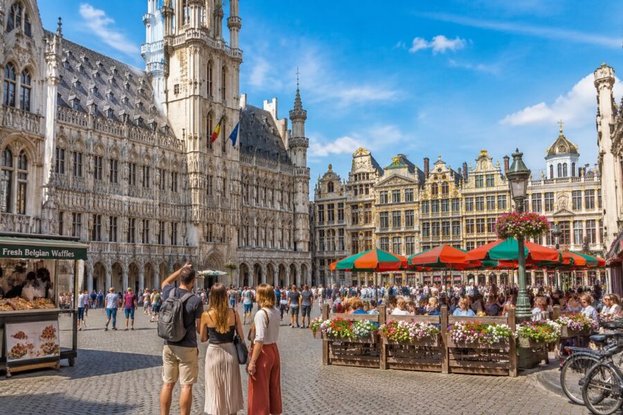 Praça central de Bruxelas com edifícios históricos, pessoas caminhando e cafés ao ar livre em um dia ensolarado.