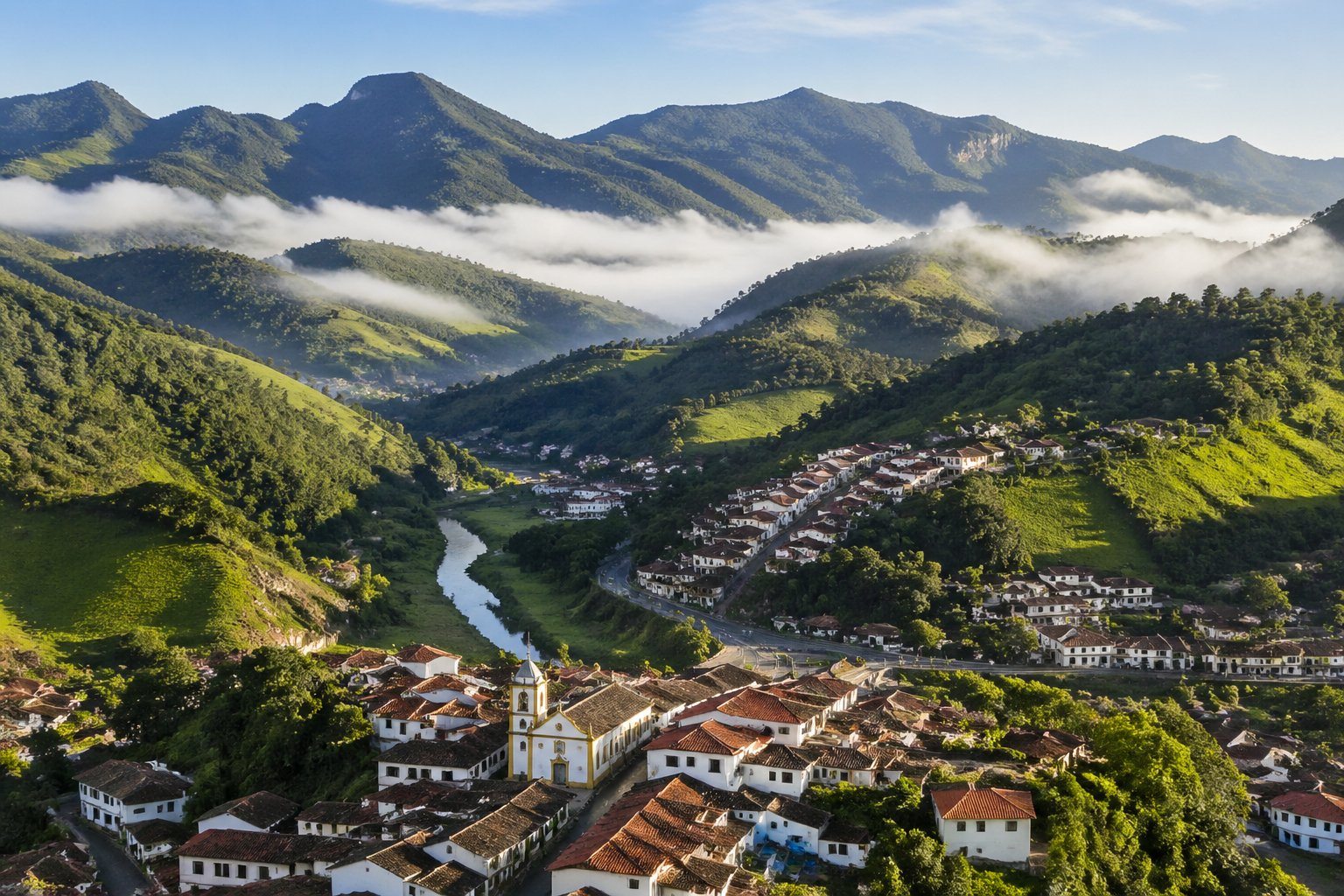 Vista panorâmica das cidades da Serra da Mantiqueira com montanhas verdes, casas com telhados vermelhos e neblina suave ao amanhecer.