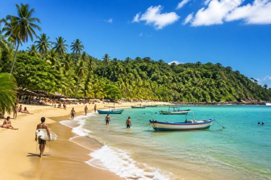 Praia tropical de Itacaré com areia dourada, água azul cristalina, coqueiros e pessoas surfando e caminhando na orla.