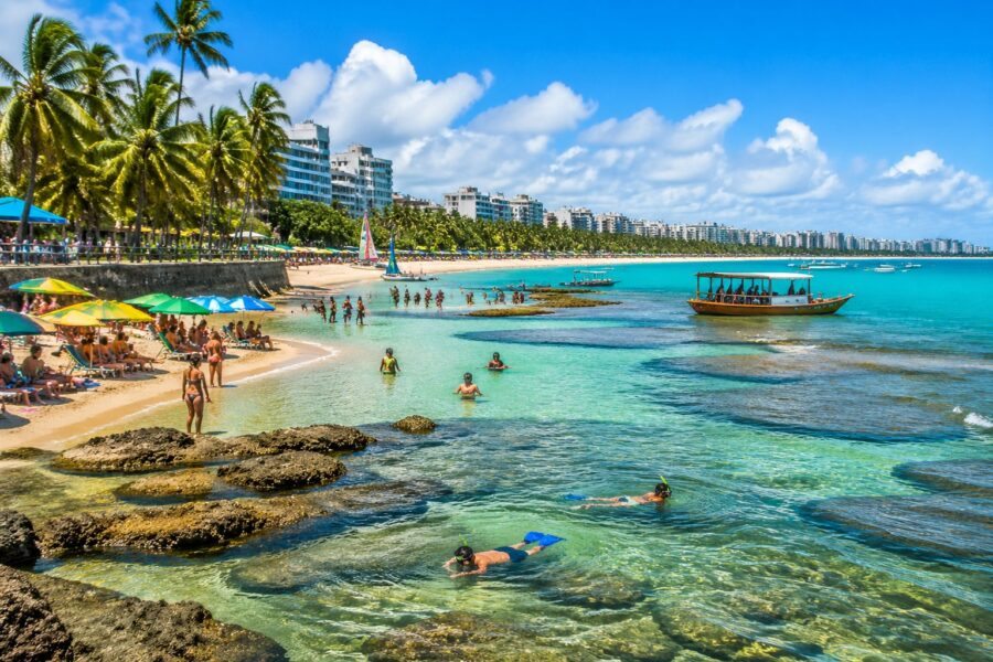 Vista panorâmica da praia de Maceió com turistas nadando, snorkelando e relaxando sob guarda-sóis coloridos, com coqueiros e prédios ao fundo.